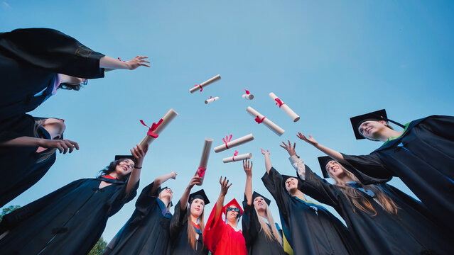 Group of students wearing graduation gowns and caps, joyfully throwing diplomas into the air against a clear blue sky, celebrating their academic achievement