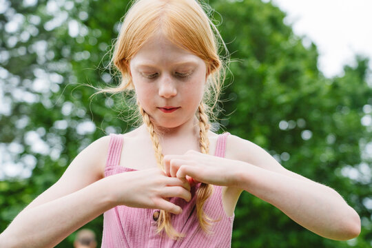 Red haired child braiding hair outdoors with focused expression