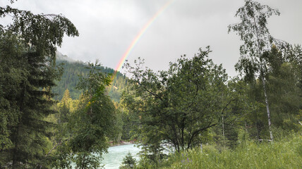 Serene natural landscape with double rainbow over river and forest. Vibrant green trees under overcast sky create visually striking and harmonious scene.