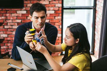 Two coworkers in modern workshop collaborate on robotics prototype at laptop. Engineering design, innovation, startup energy, and teamwork with mentoring in a creative office setting.