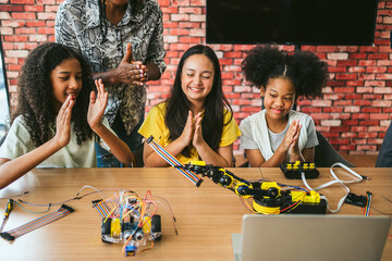 Three students celebrate successful STEM robotics test in classroom. Surrounded by laptop, coding tools, and components, they enjoy hands-on learning, education, programming, and technology workshop.