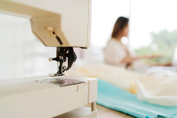 Close-up of sewing machine needle and thread on fabric in a bright atelier. Blurred seamstress in airy workshop. Tailoring, crafting, and handmade work captured in professional sewing process.