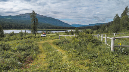 Tranquil mountain scene with a lake, lush greenery, and towering mountains. A dirt road, white vehicle, and dynamic clouds add to the serene atmosphere.
