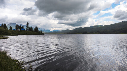 Image shows a serene mountain lake with overcast sky, calm water, mountains, clouds, trees, and a buoy. Atmosphere exudes tranquility and isolation.