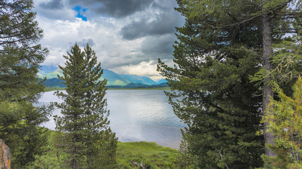 A serene landscape: calm lake, forests, mountains, fluffy clouds reflected, evergreen trees, still water contrasts with dynamic sky, tranquil beauty.