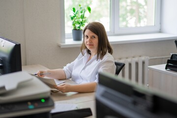 Woman in white shirt sitting at computer with paper by window