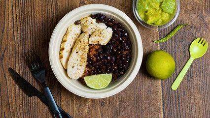 Grilled chicken breast, black beans, rice, and a slice of lime are composing a burrito bowl, served with guacamole, a lime and a chili pepper on a wooden table with black and green cutlery