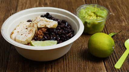 Grilled chicken breast slices, seasoned with spices, resting on a bed of black beans and rice in a biodegradable bowl, accompanied by guacamole, lime, chili pepper, and a green plastic spoon