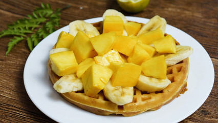 Round waffle topped with freshly cut mango and banana slices, creating a vibrant and appetizing breakfast or dessert option on a white plate