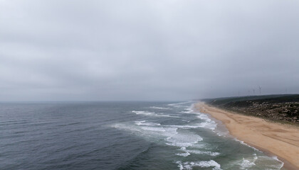 Ocean waves crashing on the sandy beach of Nazare, Portugal, on a foggy day, with wind turbines in the distance