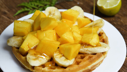 Pouring honey over a freshly made waffle, topped with vibrant slices of banana and chunks of mango, creating a sweet and irresistible breakfast delight perfect for any morning