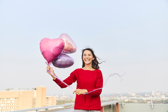 Mature woman holding heart shape balloon while standing on building terrace against clear sky