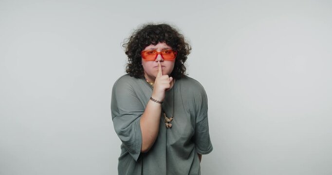 Person with curly hair wearing orange sunglasses making a shushing gesture.