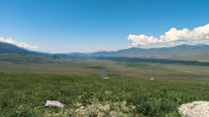 A serene grassland scene with rocks, green hills, and mountains evokes tranquility under a clear blue sky with scattered clouds.