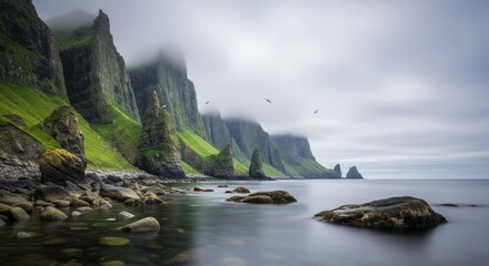 Long-exposure photograph of dramatic Karst Limestone Towers rising from the sea, showcasing a serene, misty coastal landscape with green vegetation, rocky shoreline, and