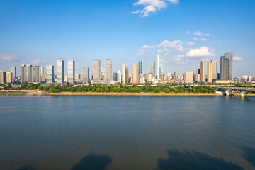 Naklejka premium Changsha City skyline in China displays modern buildings and a green riverfront reflecting in calm water under a bright blue sky.