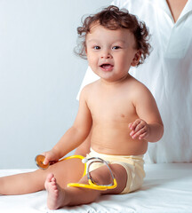 Cute curly haired toddler sitting on exam table before health check-up at hospital and playing with yellow stethoscope