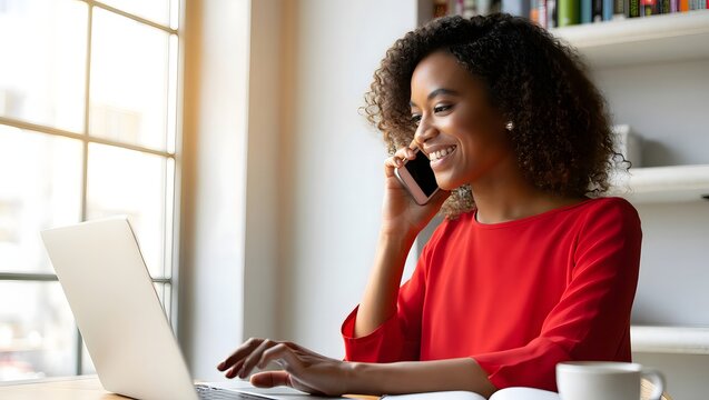 Smiling african american woman working on laptop and talking on phone at home office - Powered by Adobe