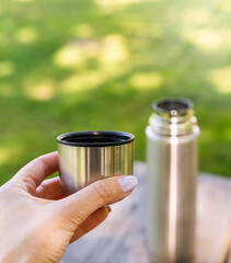 A female hand holding a stainless steel cup lid from a thermos outdoors. Concept of picnic, travel, camping, and relaxation in nature. Copy space.