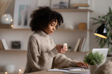 Young adult African-American business woman typing a report while sipping herbal tea, dressed in a cozy business casual sweater, working from a stylish home office with modern shelving, soft lighting,