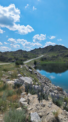 Serene outdoor scene with country road, hills, flowers, tranquil lake, and blue sky with white clouds, creating a peaceful atmosphere.