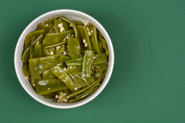 A salad of cooked green beans and garlic is served in a white bowl, placed on a green background...