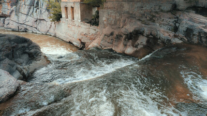 River with brown water and white foam flows through rocky channel, showcasing strong currents and turbulence. Dramatic scene from heightened perspective with trees in background.