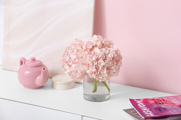 Vase with hydrangea flowers, magazines and teapot on chest of drawers near pink wall