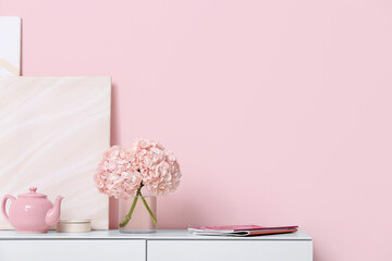 Vase with hydrangea flowers, magazines and teapot on chest of drawers near pink wall