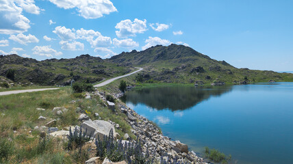 Serene landscape with calm lake, gray mountains, blue sky, and winding road. Peaceful tranquility under a bright, clear sky.