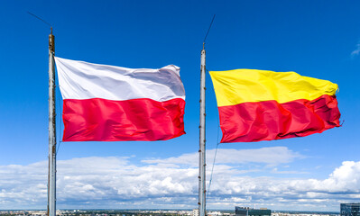 White and red Polish flag and yellow and red Warsaw flag waving on strong wind on blue sky background