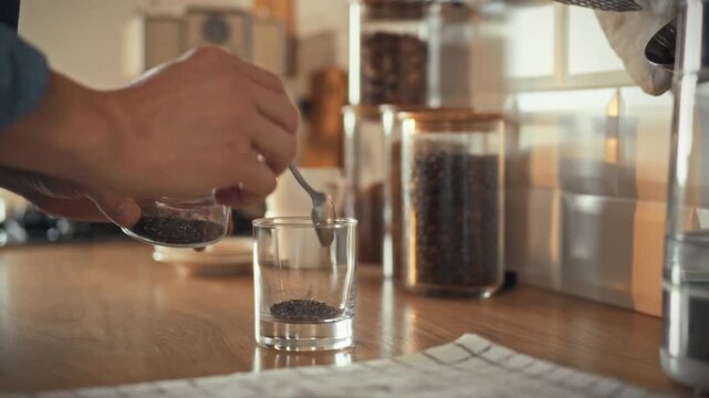 Preparing healthy breakfast man delicately places chia seeds in clear glass. A close-up shows the addition of seeds with a spoon to create healthy pudding. Balanced chia pudding for morning nutrition