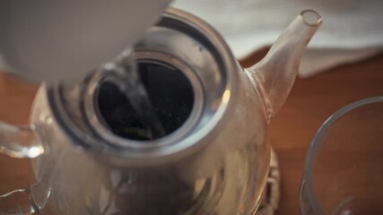Hot water is being poured into a silver teapot that contains loose leaf tea. This takes place in a cozy kitchen setting, perfect for an afternoon tea experience