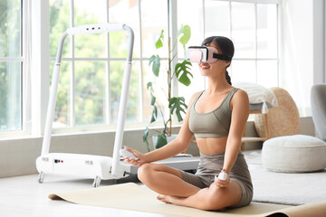 Sporty young woman in VR glasses meditating with controllers on yoga mat at home