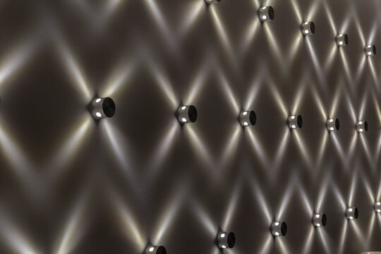 A close-up of small LED lights mounted on a wall, creating an abstract, repeating geometric pattern with crisscrossing beams of light.