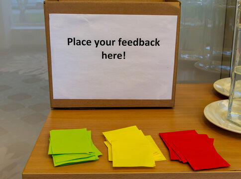 A simple cardboard box with a sign inviting people to "Place your feedback here," accompanied by colorful sticky notes. This represents collecting customer feedback and suggestions.