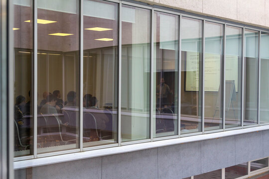 A view from outside a modern conference room with large windows, showing a group of people attending a presentation or a business meeting inside.
