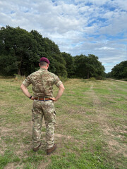 British Army soldier in maroon beret and REME stable belt, hands on waist, facing away in woodland clearing, military authority and leadership, UK armed forces.