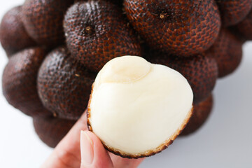 A hand holds a freshly peeled segment of salak (snake fruit), revealing the edible white flesh with unpeeled fruits in the background.