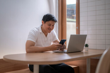 Fototapeta premium Asian man using smartphone and laptop while working remotely from a cafe