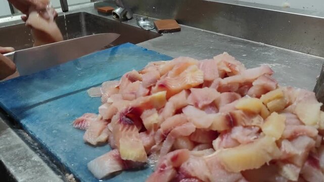 Close up of a chef&rsquo;s hands slicing fresh fish fillet on a blue cutting board in a professional kitchen. The image shows raw fish preparation for cooking, representing seafood processing, culinary work