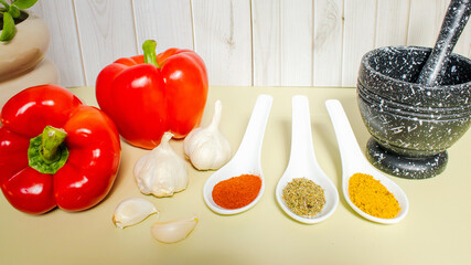 Culinary composition with spoons of bright spices for flavor, garlic in mortar and fresh vegetables on olive background. Natural ingredients, food background, healthy cooking and advertising.