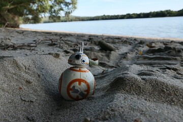 Fototapeta premium Star Wars figure of spherical rebel astromech droid BB-8 rolling in groove in sand leading from bank of Danube river. Riverside foliage, Danube river and blue skies in background. 