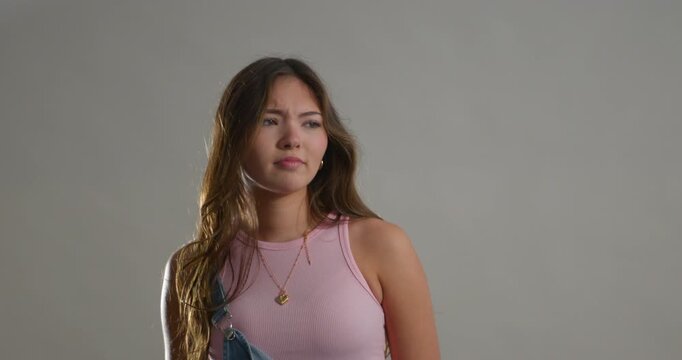 Studio video clip of a young adult female making different negative expressions. She has long brunette hair and is wearing denim dungarees with a pale pink tank top beneath.