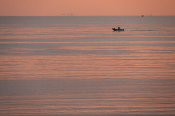 Fototapeta premium Luc-Sur-Mer, France - 08 08 2025: View of a fishing boat with a fishermen fishing with the sunrise over the sea and reflections of colorful light at sunrise