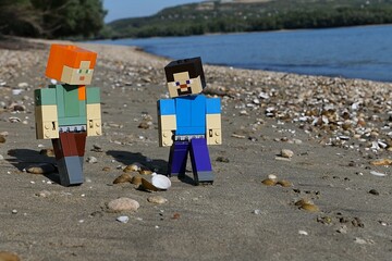 Obraz premium LEGO Minecraft action figures of Steve and Alex, examining clams and stones on sandy river Danube bank, borderline between Slovakia and Hungary. Forested opposite bank and blue skies visible. 