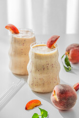 Two glasses of smoothie with peach and banana on a light background near window with tropical fruits