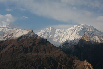Snow-capped mountain range rising above dark ridges beneath a partly cloudy sky.