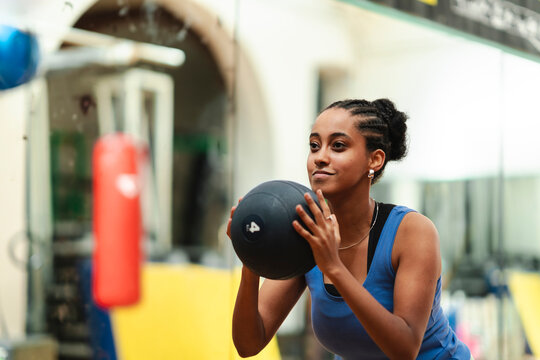 A young woman with braided hair holds a medicine ball, ready for exercise in a gym. She is focused on her fitness routine, embodying strength and healthy living.