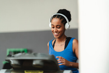 A smiling young woman with braided hair and white headphones is running on a treadmill. She is enjoying her workout at the gym, staying active and fit.